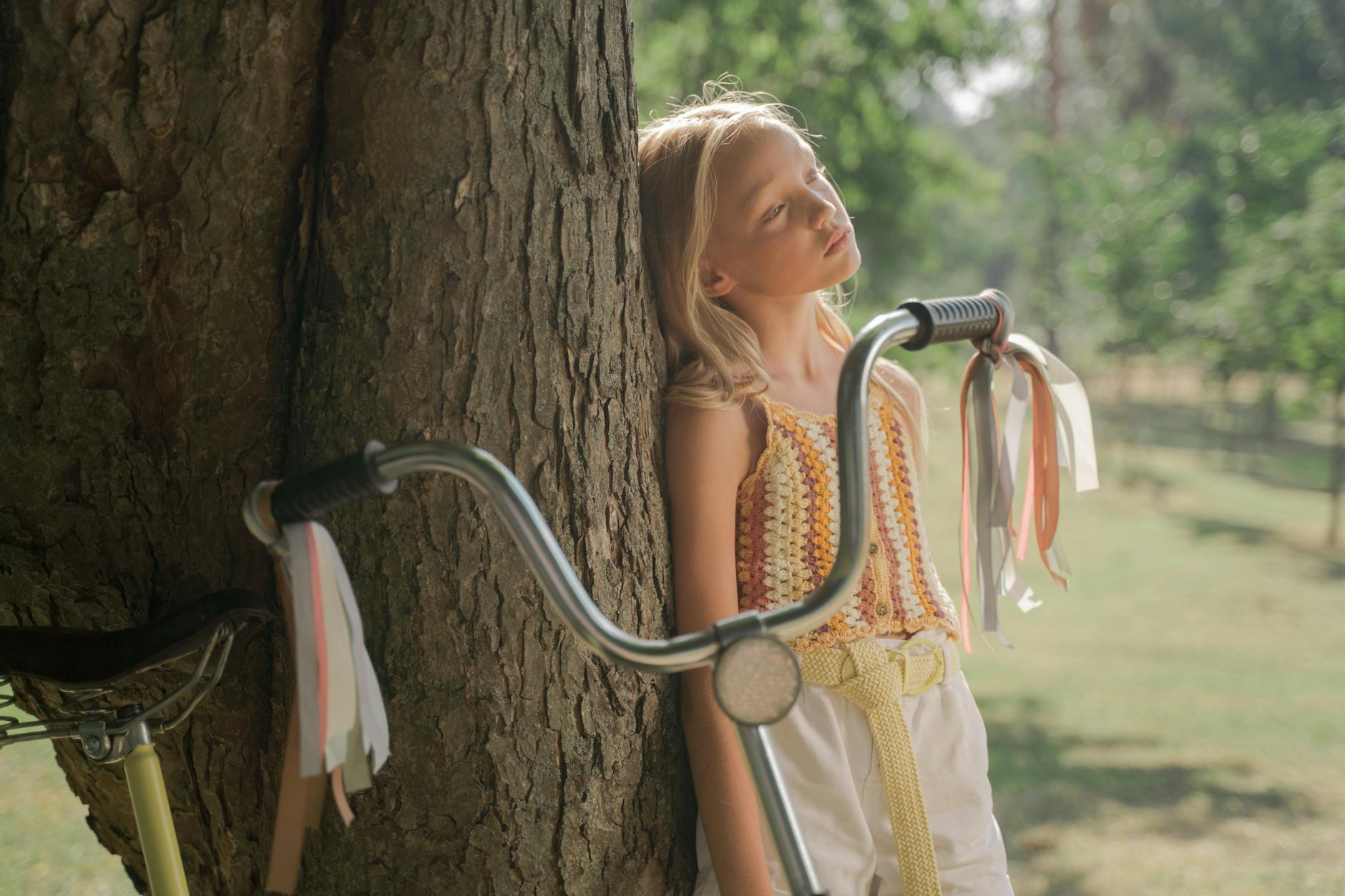 Blond girl leaning on a tree with her bicycle in a sunlit park.