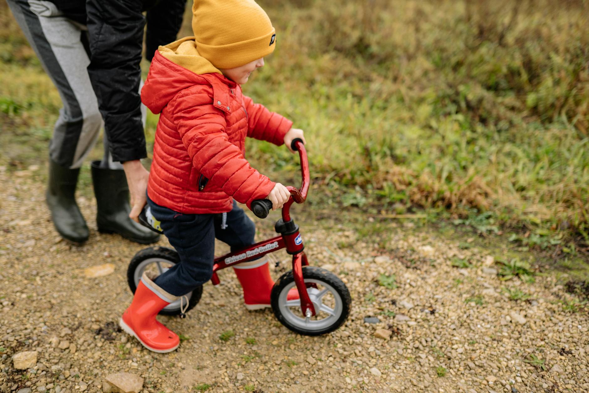 A young child in a red jacket rides a balance bike on an unpaved path outdoors.