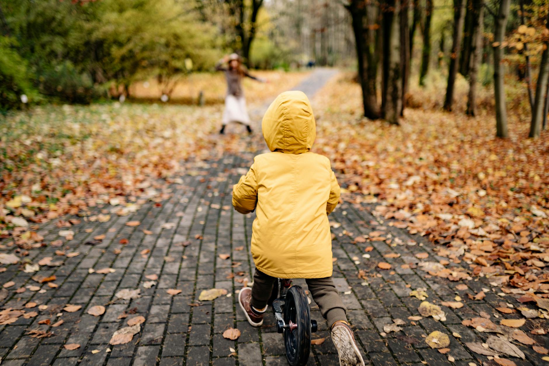 A child in a yellow jacket riding a bicycle on a leaf-covered park pathway in autumn.