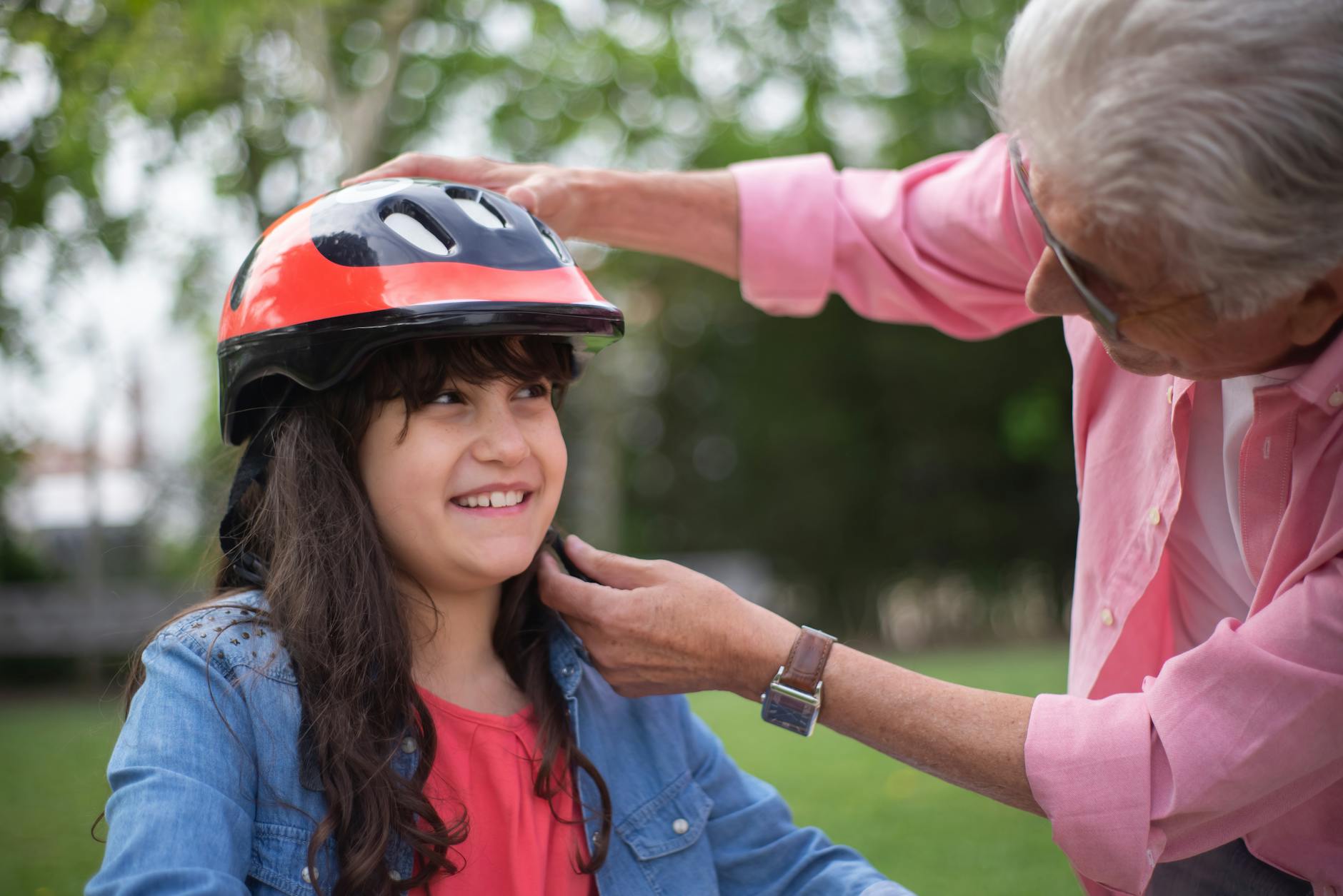 A grandfather helps his granddaughter put on a helmet, showcasing a loving family moment outdoors.