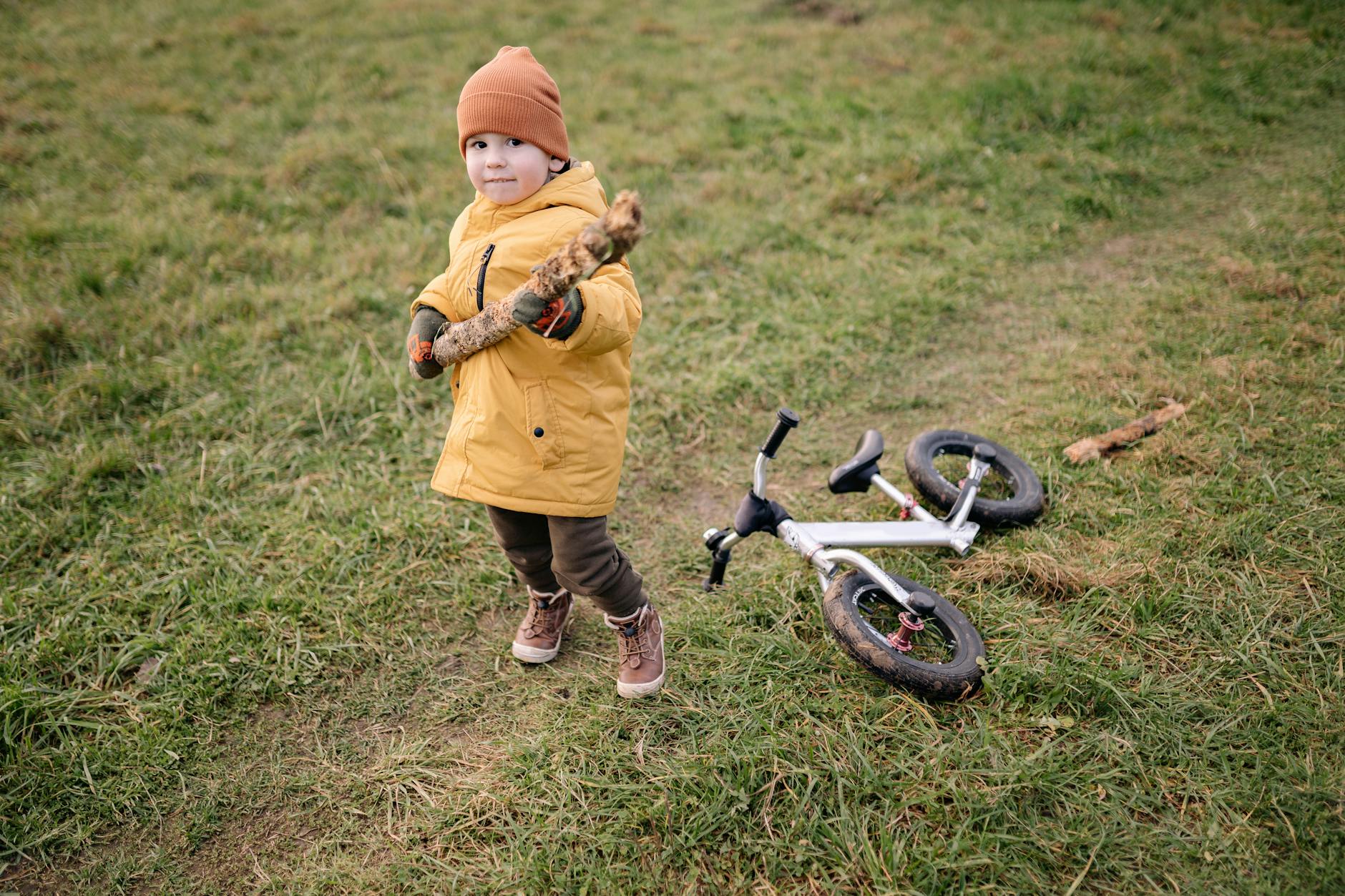 A child wearing a yellow jacket holding a stick in a grassy field near a toy bike.