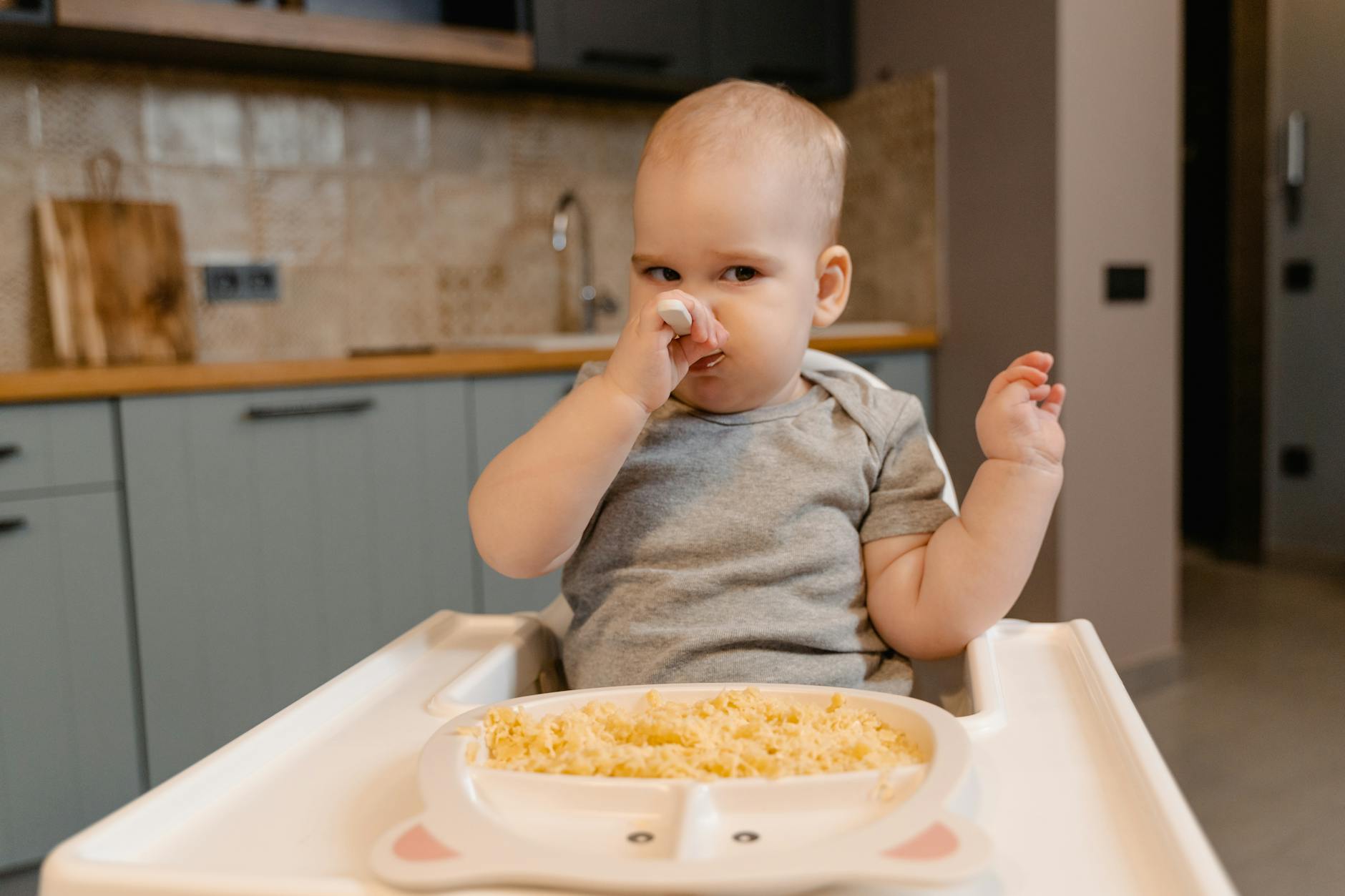 Adorable baby in onesie feeding themselves in a high chair indoors.