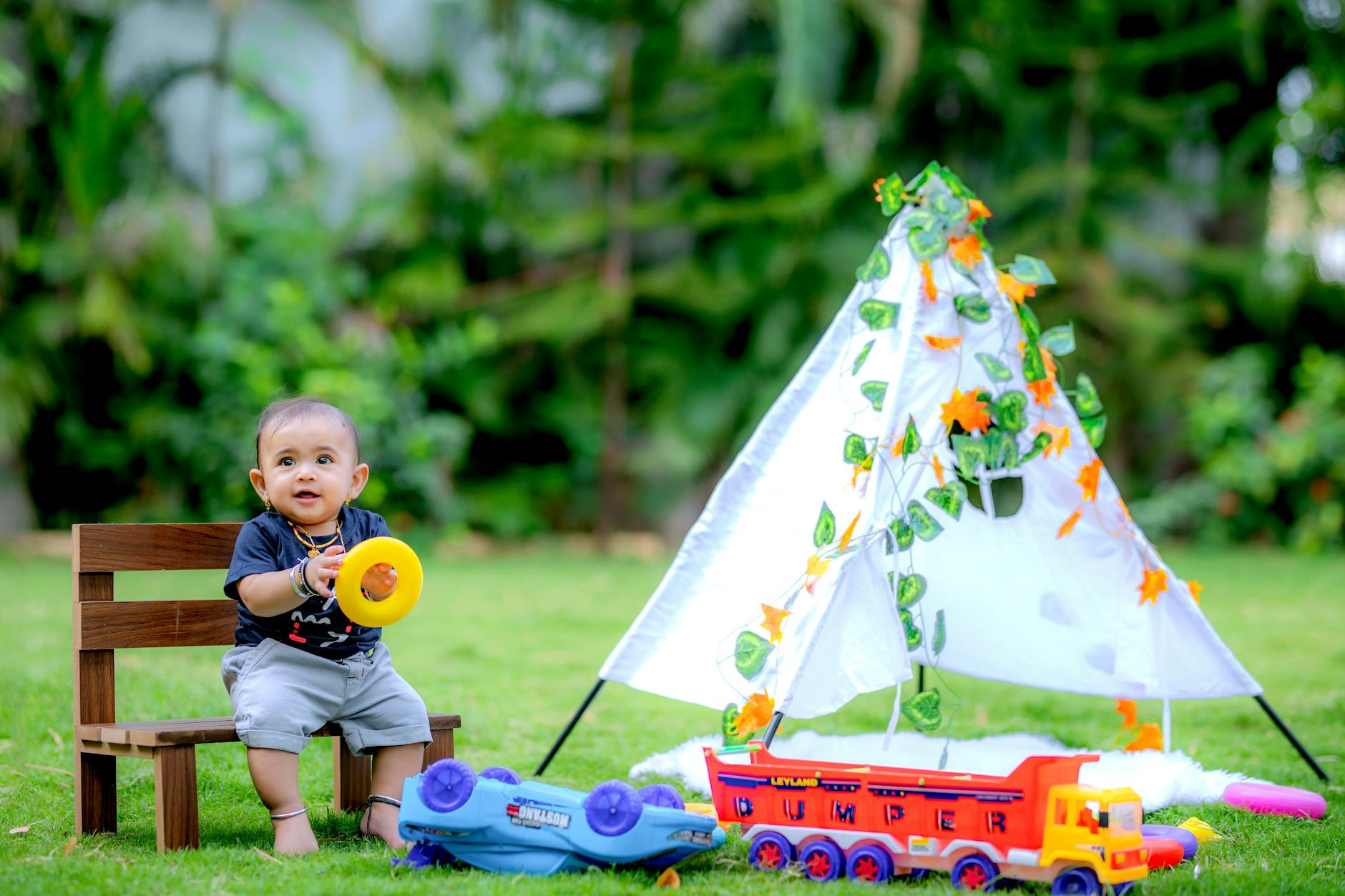Cute baby sitting on a bench with toys and a tent in a lush garden setting.