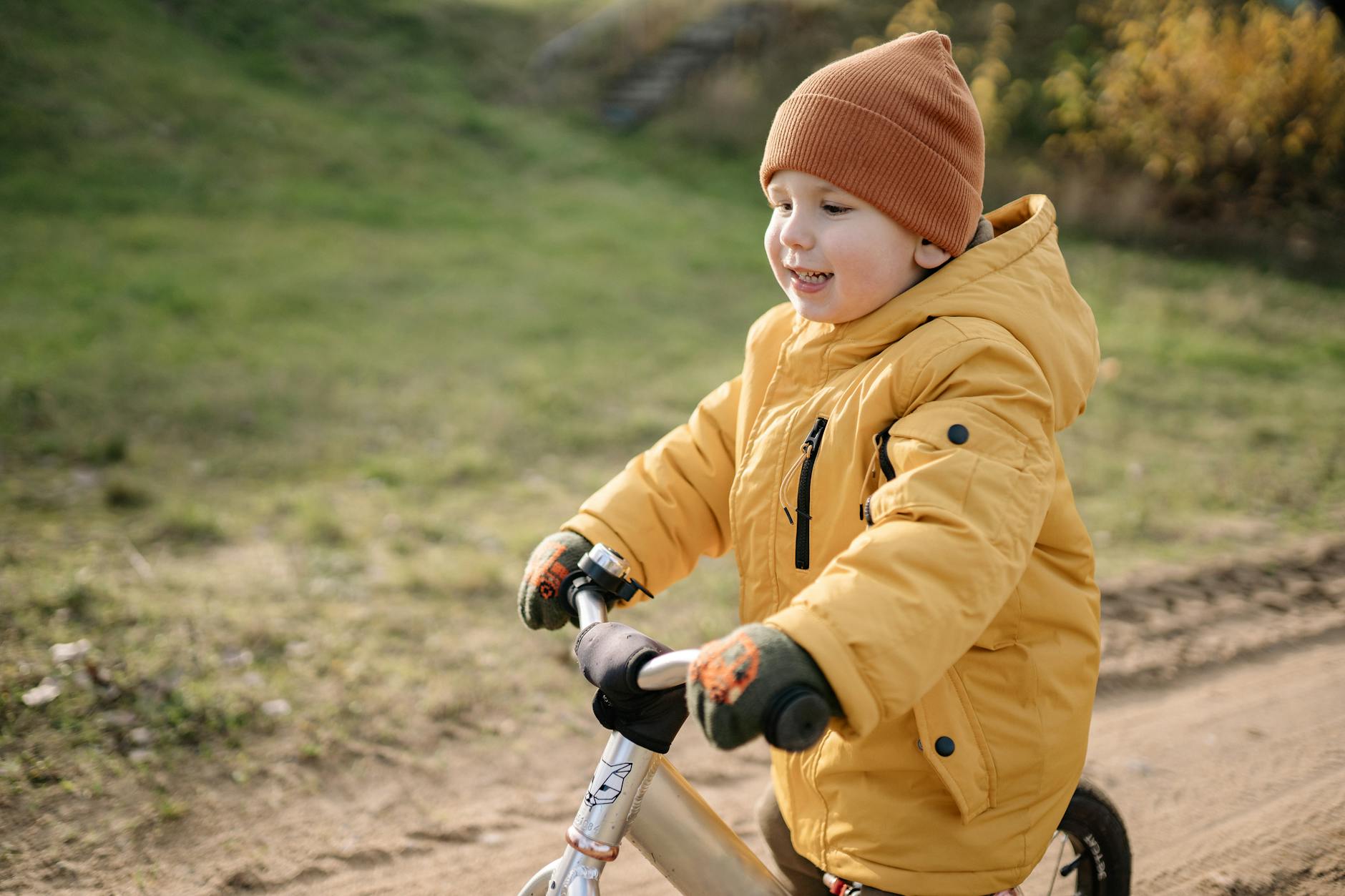 A young child wears a yellow jacket and beanie while riding a bike outdoors in fall weather.