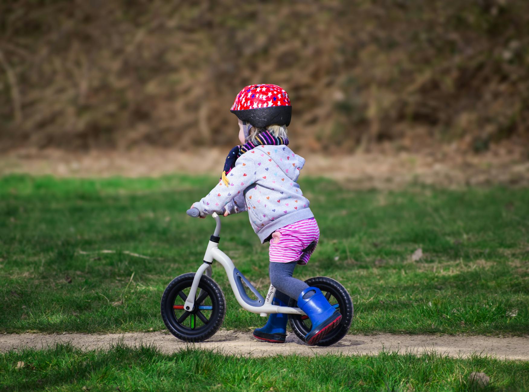 Little boy with helmet and boots riding a balance bike outdoors on a grass path.