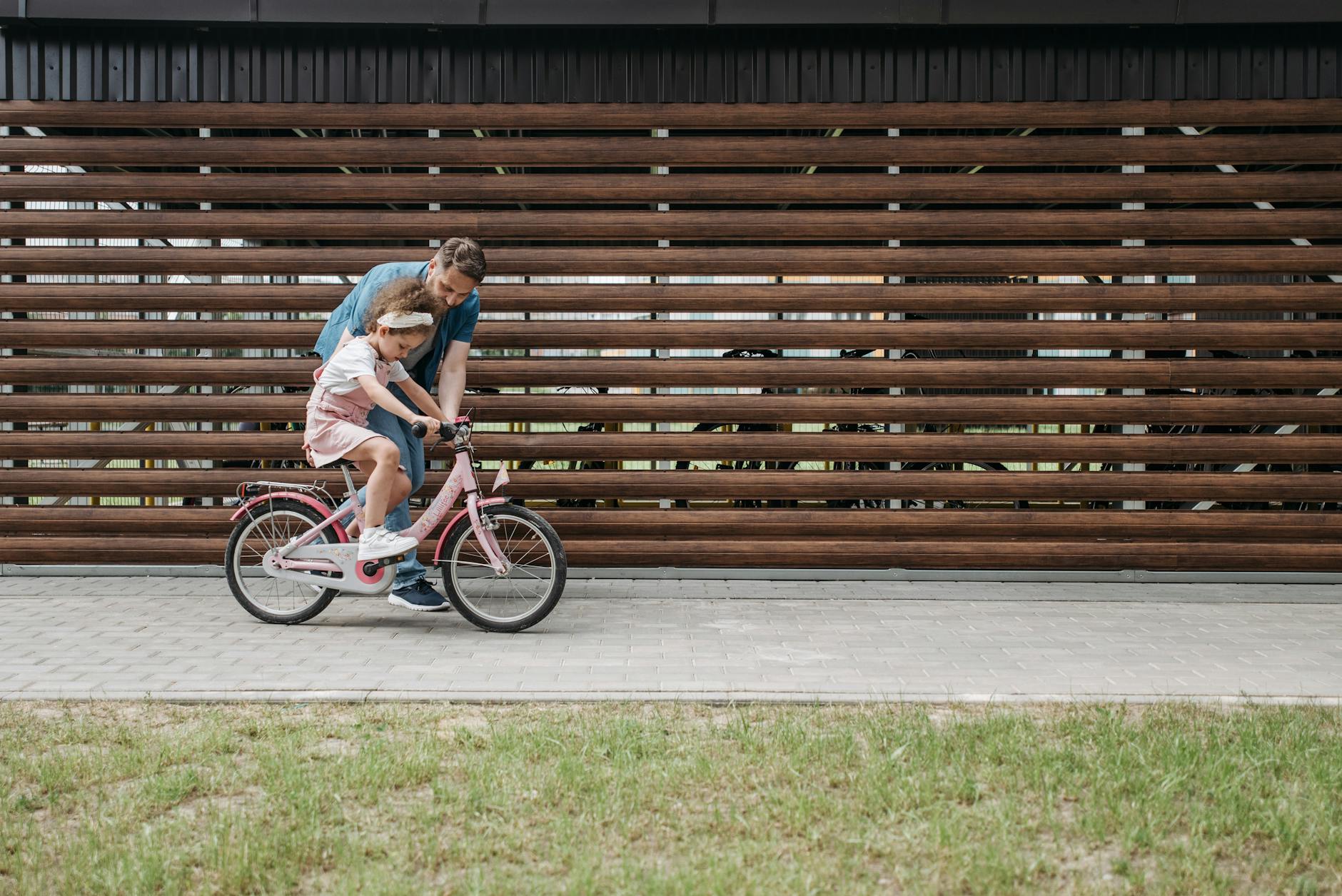 A father helps his daughter learn to ride a bike on a sunny day, fostering bonding and skill development.