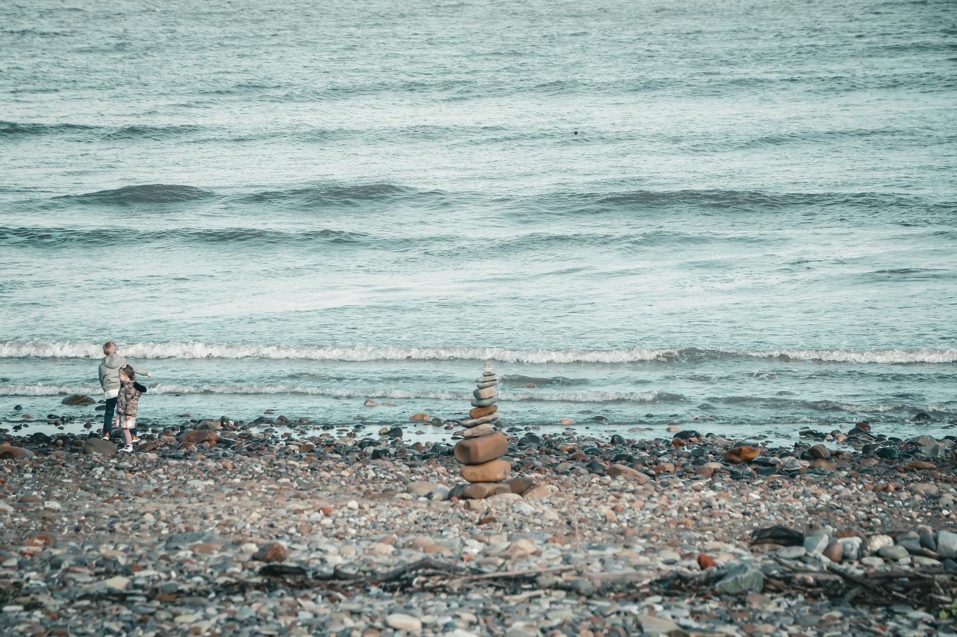 Two children playing on a pebble beach with stone cairn stacks in the foreground.