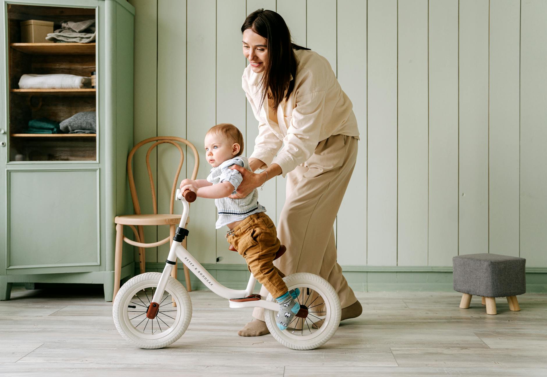 A smiling mother assists her baby learning to ride a balance bike indoors. Heartwarming moment.