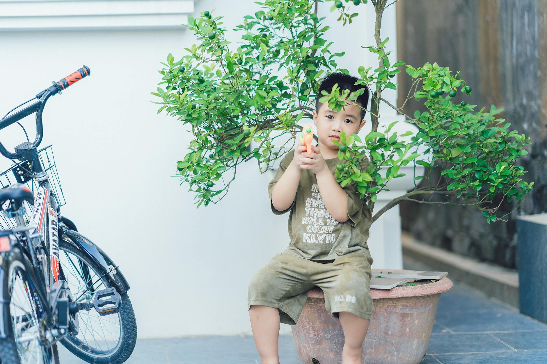 A young boy with a toy gun sits by a potted plant and bicycle outdoors.
