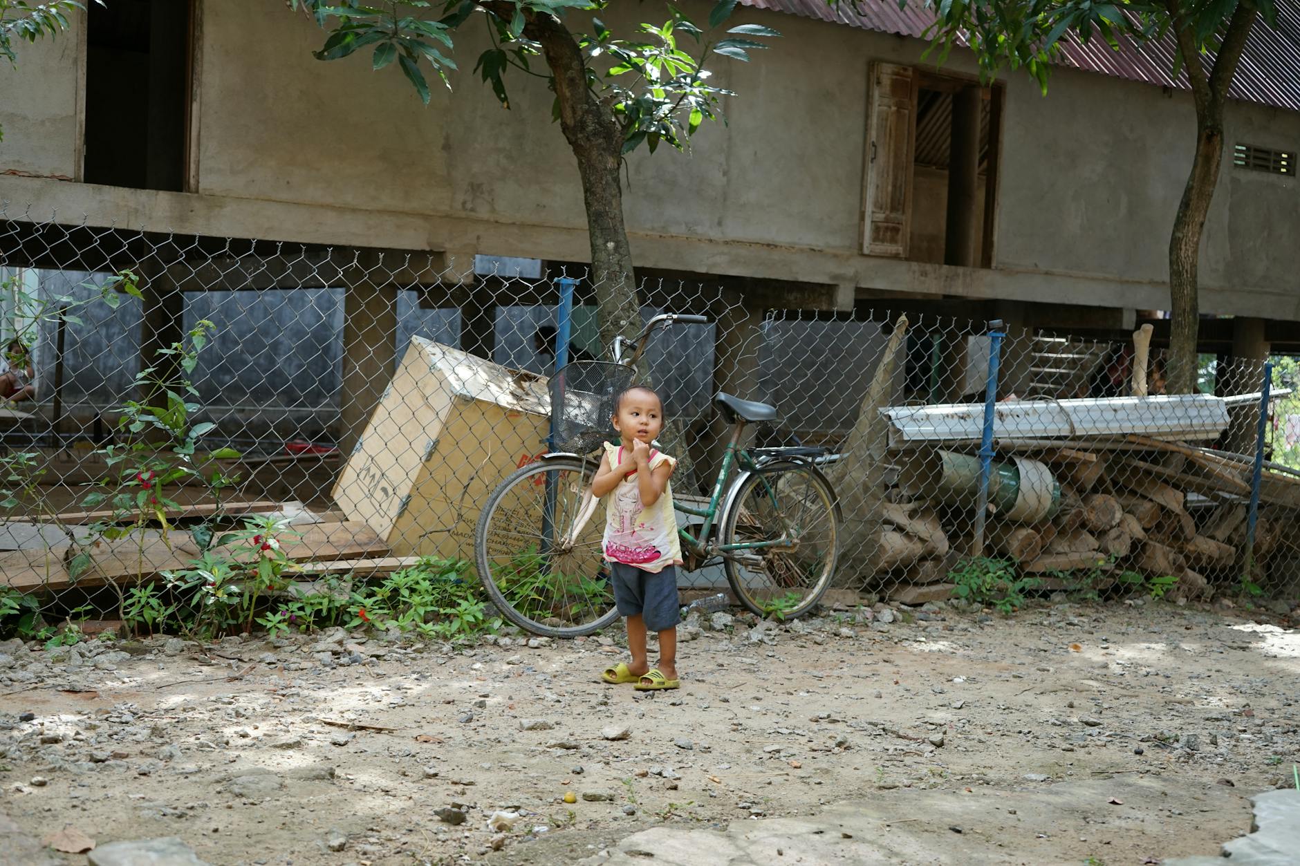 Young child standing outside in a rural setting near a bicycle and house, offering a serene scene.