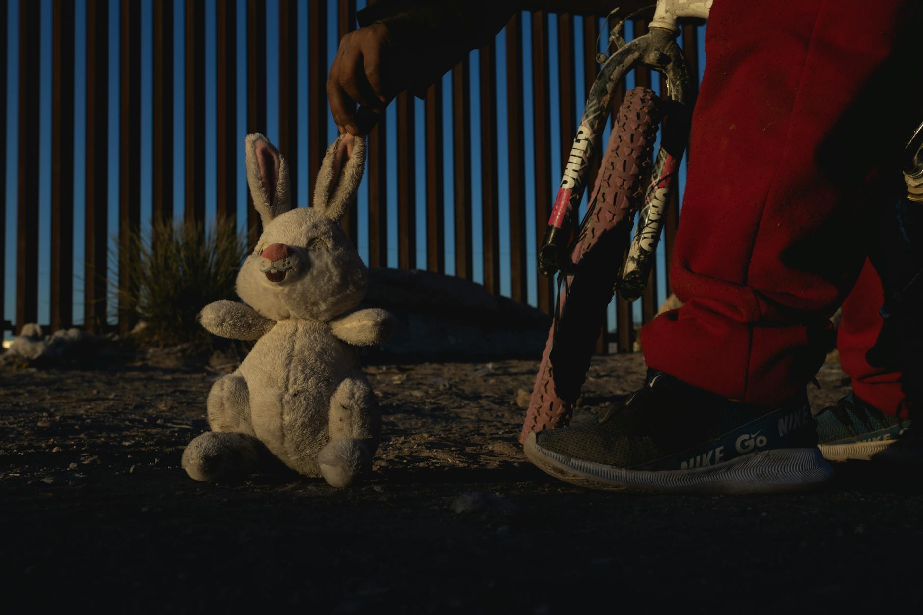 A child in red pants holds a plush bunny and a bicycle at sunset near a border fence.