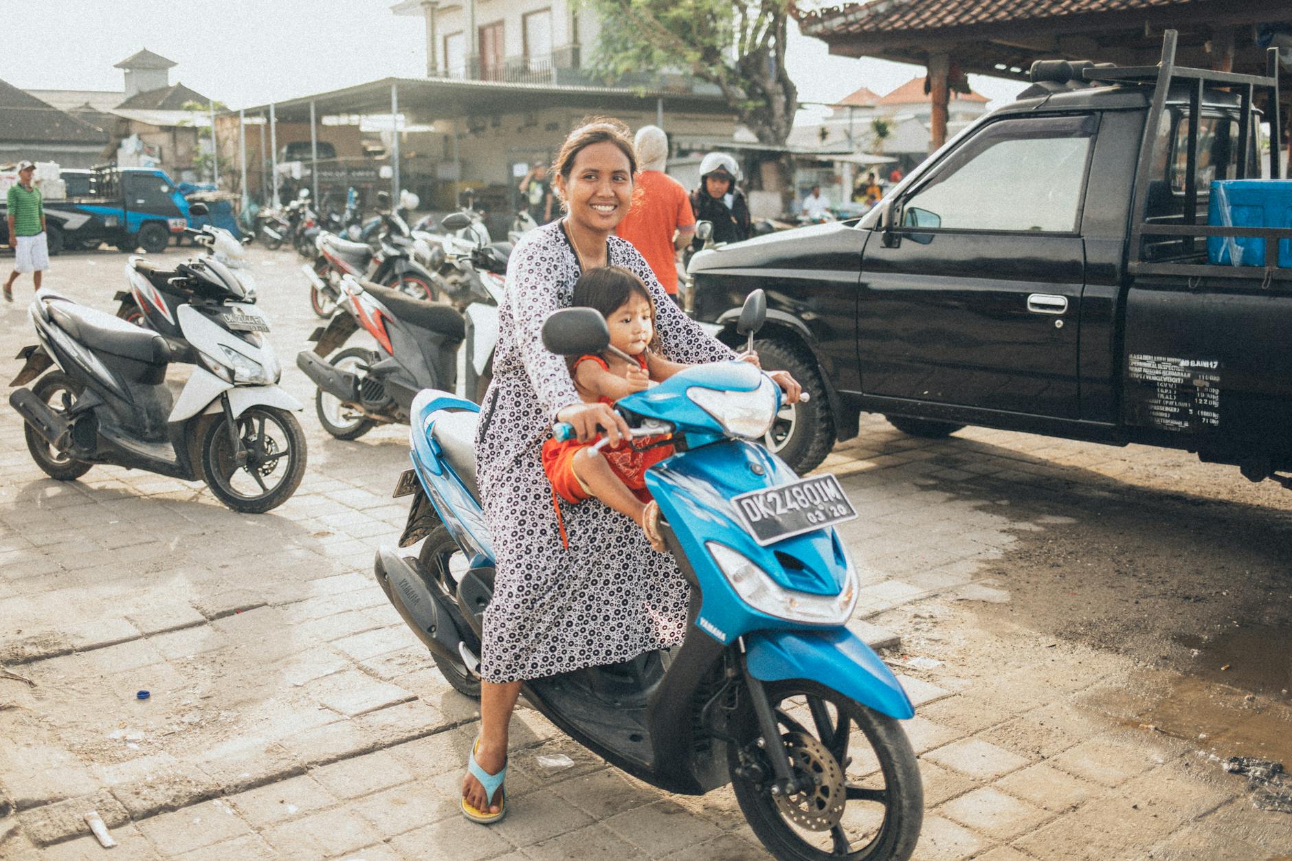 A mother and child on a blue scooter in an urban outdoor market area.