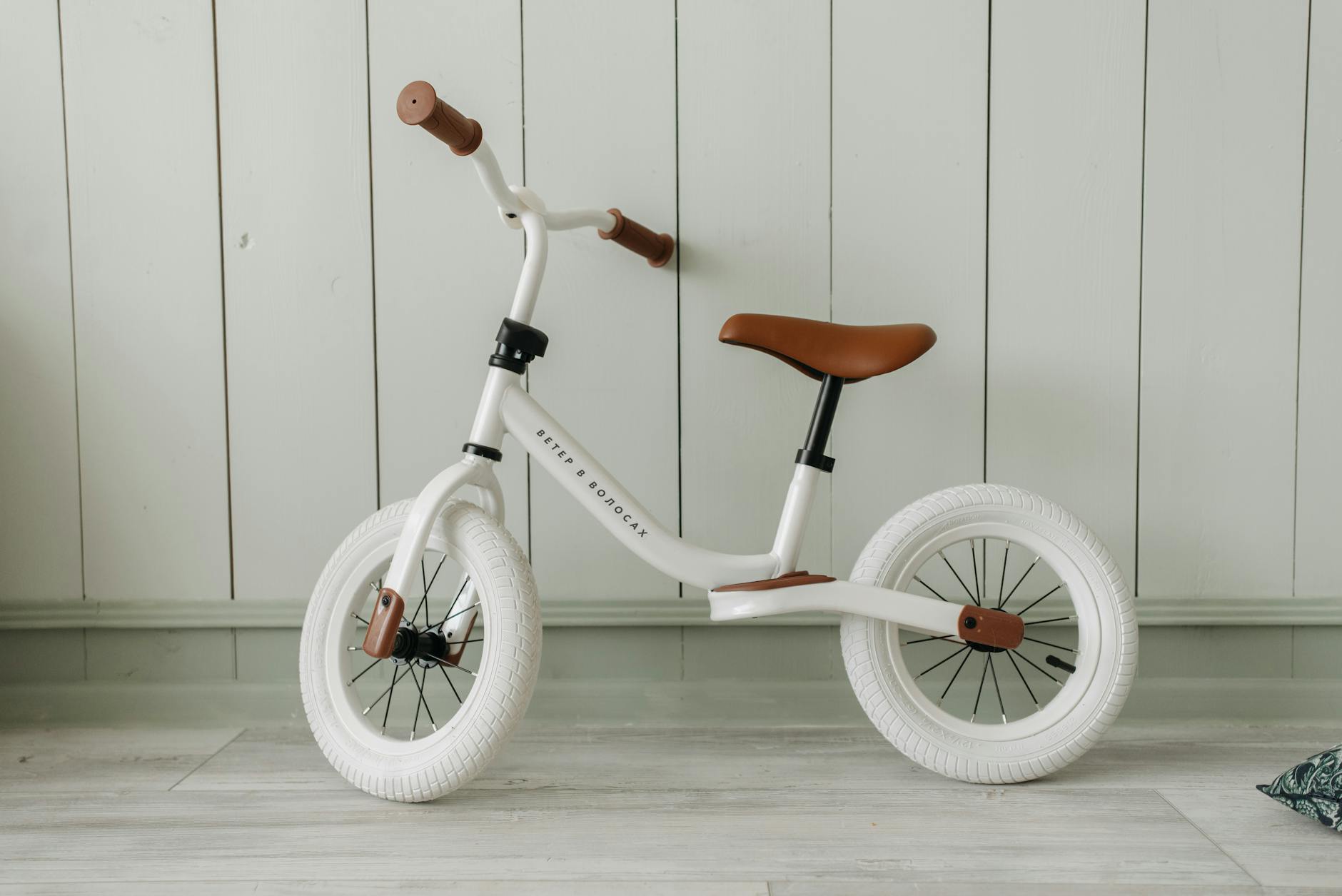 A white kids' balance bike with brown accents standing indoors against a wooden wall.